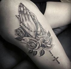 a black and white photo of a woman's thigh with a rosary, roses and praying hands tattoo