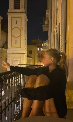 a woman is sitting on a balcony looking at the clock tower in the distance with her hand out