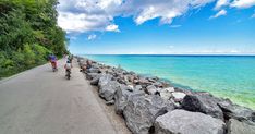 three people riding bikes on a paved road next to the ocean with large rocks in front of them