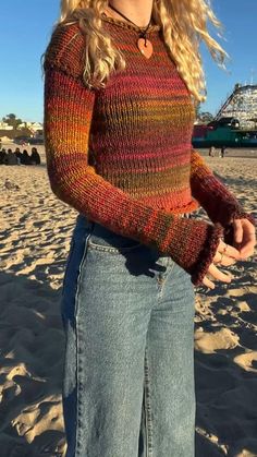 a woman standing on top of a sandy beach wearing a sweater and jeans with her hands in her pockets