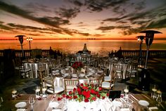 a table set up for an evening dinner by the ocean with red flowers on it