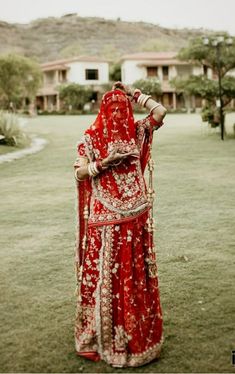 a woman in a red and gold bridal outfit standing on the grass with her hands behind her head