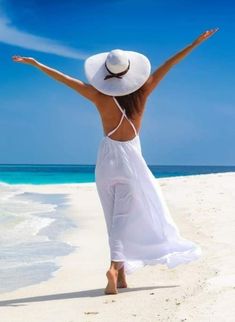 a woman in a white dress and hat on the beach with her arms outstretched up