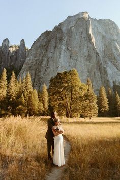 a bride and groom standing in front of a mountain with tall grass on the ground