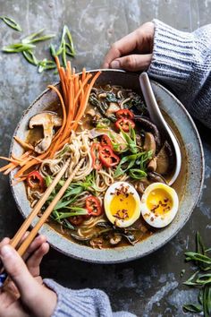 two hands holding chopsticks over a bowl of noodles and vegetables with hard boiled eggs