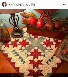 a table topped with a red and green quilt next to a vase filled with apples