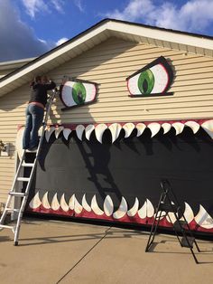 a man on a ladder painting a fake monster's face onto the side of a house