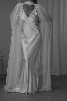 Bride wears a simple slip dress created with silk satin inside a bridal shop. her hair is up in a bridal pony. she styles her wedding dress with a silk chiffon cape. the cape has an ethereal look with draping along the shoulders and an elegant necktie accent. the look is ethereal and luxurious for her modern wedding. Scarf For Wedding Dress, Wedding Dresses With Shawls, Sheer Top Over Wedding Dress, Sheer Bridal Cape, Bridal Shaw, Wedding Gown Cape, Wedding Dress With Cape Sleeves, Wedding Dress Cape, Wedding Dress With Cape