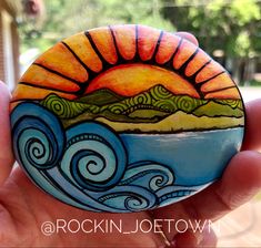 a hand holding a painted rock with the sun setting over a body of water and mountains