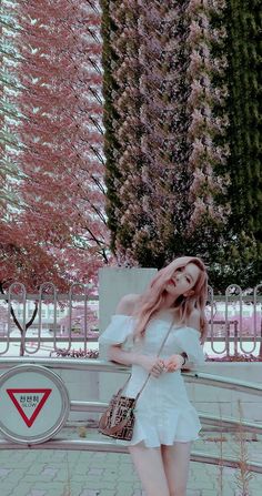 a woman in white dress holding a purse and standing next to a tree with pink flowers