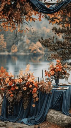 an outdoor table set up with flowers and greenery on the table, next to a lake