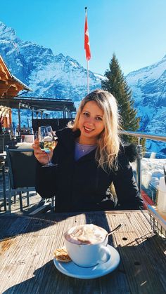 a woman sitting at a table with a glass of wine in front of her and mountains behind her