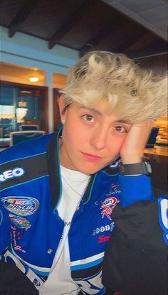 a young man with blonde hair wearing a blue and black racing jacket sitting in front of a table