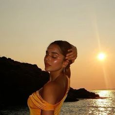 a woman standing in front of the ocean with her hands on her head as the sun sets