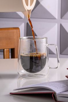 a person pours coffee into a glass mug on a table with an open book