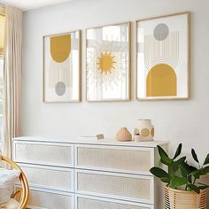 two paintings on the wall above a dresser in a room with white furniture and plants