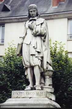 a statue of a man holding a book in front of a building with trees and bushes