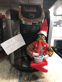 an elf is sitting next to a coffee machine with a sign on it that says elf refreshment