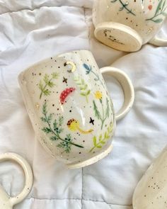 three coffee mugs with designs on them sitting on a white bed sheet, one is upside down and the other two are empty