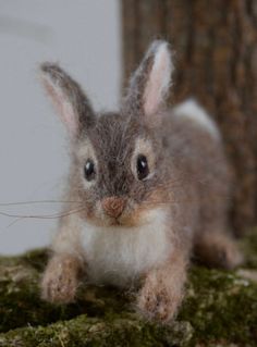 a needle - felted bunny rabbit sitting on mossy ground next to a tree