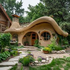 Discover a whimsical cob house featuring a curvaceous roof, warm beige walls, and rounded windows. The wooden arched door and stone accents enhance its rustic charm. A stone pathway winds through a lush garden filled with vibrant flowers and greenery, creating a serene and enchanting atmosphere. This cob house seamlessly blends natural beauty with unique architectural design, offering a picturesque and tranquil retreat. Sustainable Cottage, Whimsical Tiny House, Cob Castle, Small Cob House, Round Cottage, Cobb Cottage, Cob Cottage Exterior, Adobe Houses, Round Cob House