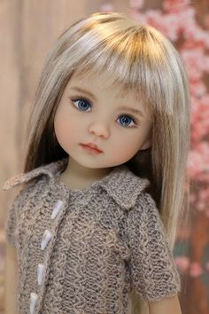 a doll with blonde hair and blue eyes is wearing a knitted sweater in front of a cherry blossom tree