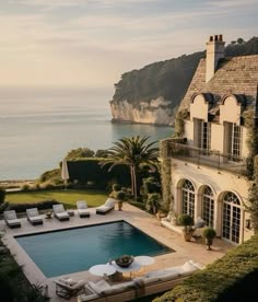 an outdoor swimming pool next to a large house near the ocean with lounge chairs around it