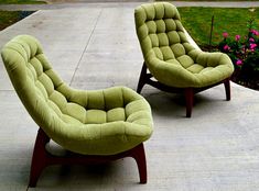 two green chairs sitting on top of a cement floor next to flowers and bushes in front of a house