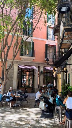 people are sitting at tables in an outdoor cafe area with trees and buildings behind them