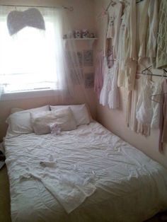 an unmade bed in front of a window with clothes hanging on the wall behind it