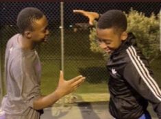 two young men standing next to each other near a fence and one pointing at something