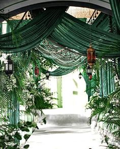 an archway covered with green curtains and hanging lanterns in the middle of a walkway surrounded by greenery