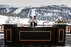 two men standing behind a bar in front of a snowy mountainside area with flowers and candles on it