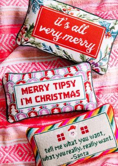 three cross stitch christmas pillows on a pink and white checkered blanket with the words merry tipsy i'm christmas