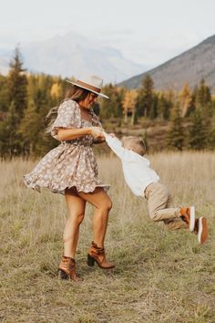 a woman in a dress and hat playing with a small child on the grass near mountains