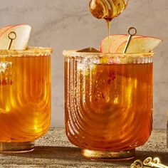 an apple cider being poured into two glasses filled with apples and gold spoons