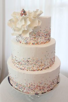 a three tiered cake with sprinkles and a white flower on top