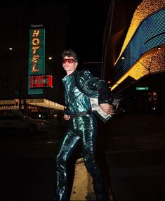 a man in shiny pants and sunglasses walking down the street at night with his hand on his hip