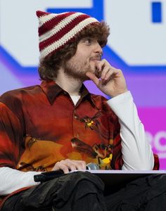 a man with a hat on sitting in front of a laptop computer and holding his hand to his chin