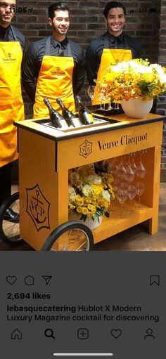 three men in yellow aprons stand behind a cart with bottles and glasses on it