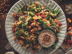 a white plate topped with pasta salad next to bowls of vegetables and sauces on a wooden table