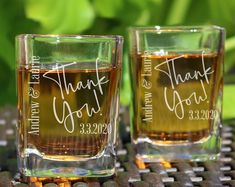 two shot glasses with names on them sitting on a table next to each other and green leaves in the background