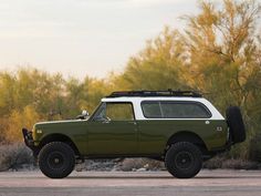 a green truck parked on the side of a road next to some bushes and trees