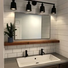 a bathroom sink under a large mirror with lights on the wall and a potted plant next to it