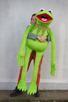 a woman is hugging a large green frog mascot in front of a white wall and grass area