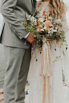 a bride and groom standing next to each other on the beach with flowers in their bouquets