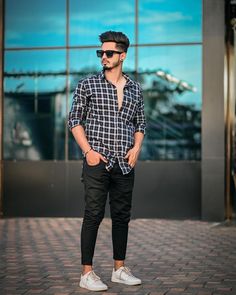 a man standing in front of a building wearing black pants and a plaid shirt with white sneakers