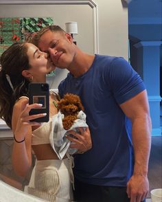 a man and woman kissing while holding a teddy bear in front of a bathroom mirror