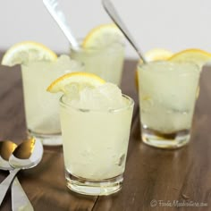 three glasses filled with lemonade and ice on top of a wooden table next to spoons