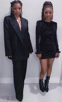 two women standing next to each other in front of a white wall wearing black clothing